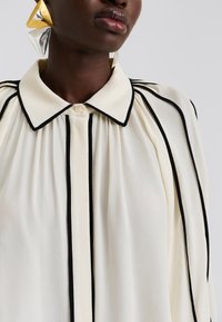 Κρεμ μπλούζα με μαύρες κάθετες ρίγες, με δομημένο γιακά και κουμπώματα. Το ύφασμα είναι λείο με ελαφριά γυαλάδα.