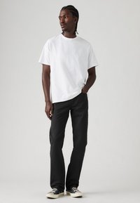 Witte t-shirt met korte mouwen, gecombineerd met zwarte broek en zwarte sneakers. De stof lijkt glad, met een ontspannen pasvorm en minimalistisch design.