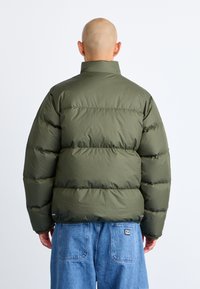 Olive green puffer jacket, featuring a high collar, ribbed cuffs, and vertical quilted design. Worn over light blue denim pants.