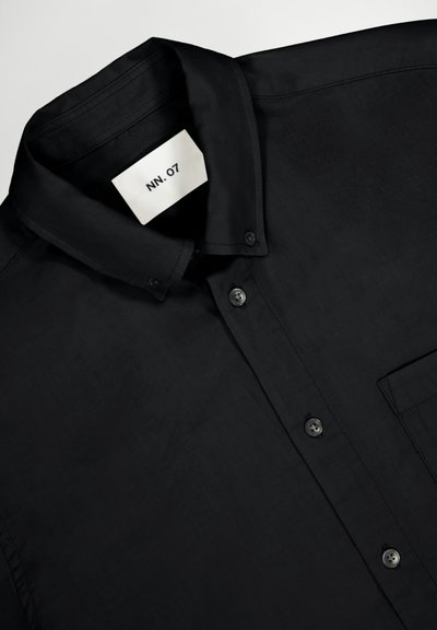 Chemise noire à boutons, fabriquée en tissu lisse, dotée d'un col structuré, de manches longues, d'une poche avant et de boutons noirs. L'étiquette indique "NN. 07."
