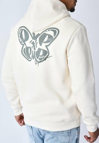 Sudadera con capucha color crema, con un gran gráfico de mariposa gris en la parte posterior y detalles de firma; puños y dobladillo acanalados; textura de tela suave.
