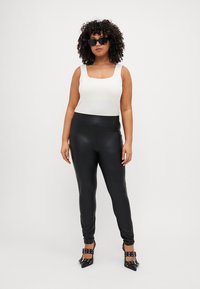 Fekete műbőr leggings magas derékkal, egy testhez álló fehér ujjatlan felsővel párosítva. A modellen szögletes fekete napszemüveg és hegyes sarkú cipők vannak.