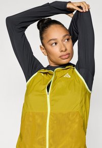 Ζακέτα με φερμουάρ, με κίτρινο κορμό και μαύρα μακριά μανίκια, κατασκευασμένη από ελαφρύ, υφή ύφασμα και νεον πράσινες πινελιές κατά μήκος των φερμουάρ.