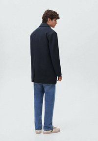 Joven con cabello rizado que lleva un abrigo azul marino, jeans azules y zapatillas color beige, de pie y mirando hacia un lado contra un fondo sencillo.
