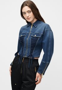 Νεαρή γυναίκα με μακριά καστανά μαλλιά που φοράει εφαρμοστό μπλε τζιν μπουφάν με δέσιμο στο μπροστινό μέρος και μαύρο παντελόνι με χρυσές λεπτομέρειες φερμουάρ.