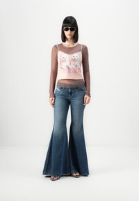 Kvinne i utsvingte blå jeans, rosa topp med kattemotiv over gjennomsiktig langermet skjorte, svarte solbriller og svarte flip-flops.