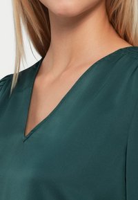 Zelena bluzica s V-izrezom, izrađena od glatkog materijala. Sadrži suptilne nabore na ramenima za dodatnu teksturu i detalj.