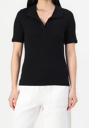 Vrouw draagt een aansluitend zwart polohemd met korte mouwen gecombineerd met witte broek met hoge taille tegen een effen achtergrond.