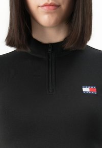 Vrouw in een zwart zip-up shirt met een klein Tommy Jeans-logo op de borst, met recht donker schouderlang haar.