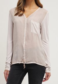 Chemise beige clair à manches longues avec col en V, boutons sur le devant, une poche poitrine unique et un ourlet à cordon de serrage. Le tissu semble doux et léger.