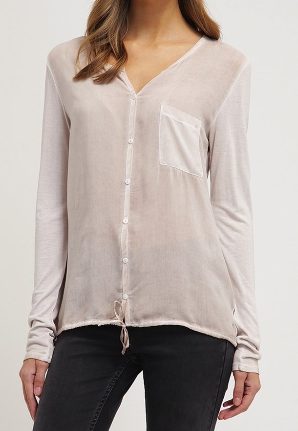 Chemise beige clair à manches longues avec col en V, boutons sur le devant, une poche poitrine unique et un ourlet à cordon de serrage. Le tissu semble doux et léger.
