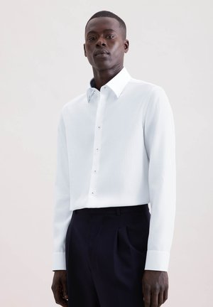 Mann trägt ein weißes Hemd mit Knöpfen und dunkle Hose, steht vor einem schlichten hellen Hintergrund, blickt nach vorne.