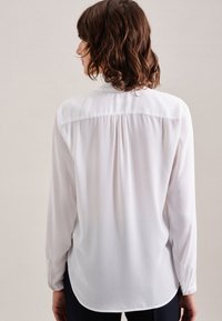 Weiße Bluse mit langen Ärmeln, glatter Textur und dezenten Falten am Rücken, mit einem Kragen und Knopfmanschetten.
