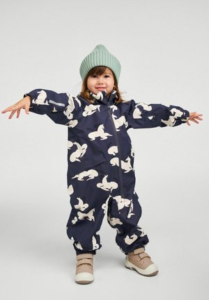 Maluch ubrany w granatowy kombinezon z białymi nadrukami zwierząt, miętowo-zieloną dzianą czapkę i beżowe buty, stojący z rozpostartymi ramionami na jednolitym tle.