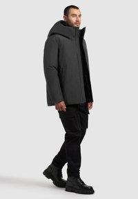 Black insulated jacket with a high collar and large hood, featuring a front zipper and side pockets, worn over black cargo pants and boots.