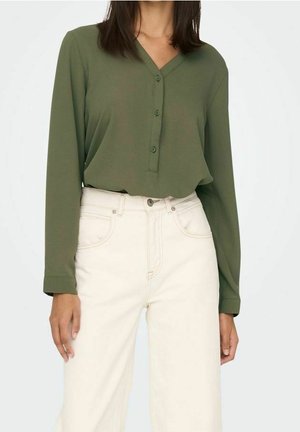 Femme portant un chemisier vert olive à manches longues avec des boutons et un pantalon taille haute écru sur un fond uni.