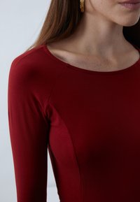 Burgundy long-sleeve top with a rounded neckline and smooth texture. Close-up shows shoulder seam details and subtle stretch in fabric.
