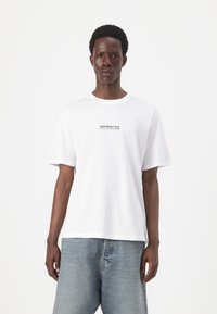 T-shirt bianca in cotone con maniche corte, scollatura rotonda e scritta nera "BRILLA FORTE CON SOGNI METALLICI" sul petto.