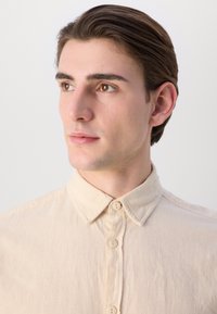 Beige, button-up shirt with a soft texture and a classic collar design. Features visible stitching and a relaxed fit.