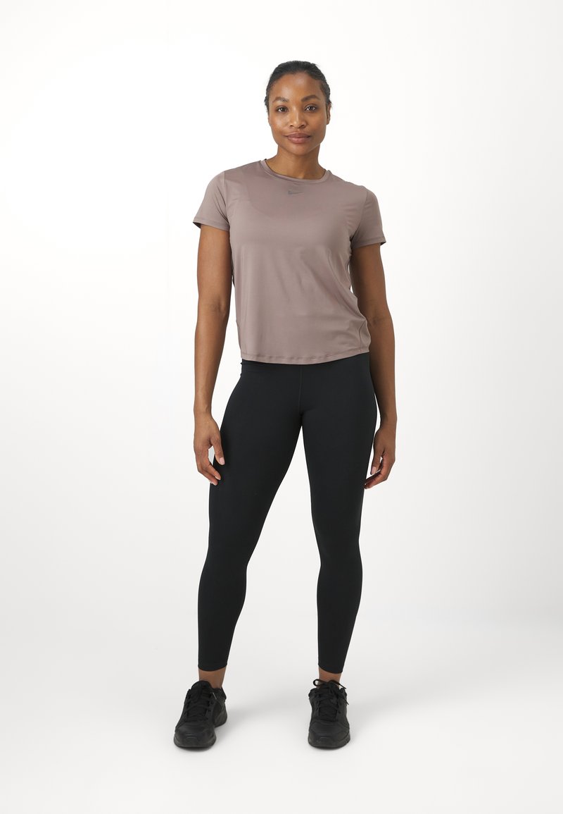 Vrouw staat, draagt een mauve sporttop met korte mouwen, zwarte legging en zwarte sneakers tegen een witte achtergrond.