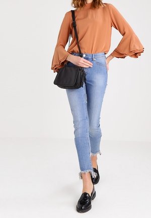 Langärmlige rostfarbene Bluse mit Rüschensaum, hellblaue destromisierte Skinny-Jeans mit fransigem Saum und schwarze Umhängetasche. Schwarze Slipper.