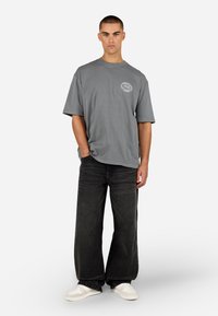 Pelēks pārprodukta t-krekls ar apaļu grafisko logo uz krūtīm, kombinēts ar platām melnām biksēm un baltām tenisa kedām, uz viendabīga fona.