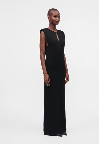 Grande personne aux cheveux courts portant une longue robe noire sans manches avec une petite ouverture frontale et un bouton doré, debout sur un fond blanc.