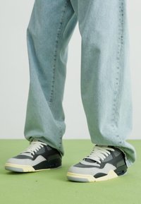 Grå og sorte sneakers med hvide snørebånd; grå tekstiloverdel og gummisåler; parret med lyseblå jeans på et grønt gulv.