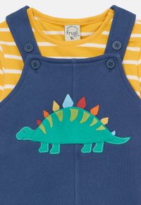 Modra bombažna dungaree obleka s podloženo rumeno črtasto majico. Ponuja vezenje zelenega dinozavra z barvitimi bodicami. Prilagodljivi gumbi na naramnicah.