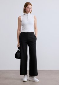 Λευκό αμάνικο τοπ με φερμουάρ στο γιακά, σε συνδυασμό με μαύρο παντελόνι φαρδύ στο τελείωμα με ζώνη που φέρει το λογότυπο Calvin Klein. Λευκά αθλητικά παπούτσια και μια μικρή μαύρη τσάντα.