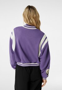 Lilla cropped sweatshirt med hvide striber på ærmerne og kraven. Har et rundt logo på venstre ærme og en struktureret, blød stof.