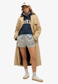Beige trenchcoat, marineblauwe hoodie met witte tekst, gelaagd blauw geruite shirt, grijze shorts, witte sneakers en grijze sokken.