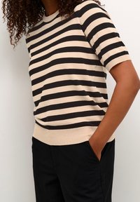 Mujer con cabello rizado que lleva un suéter de manga corta a rayas horizontales beige y negro y pantalones negros, con una mano en el bolsillo, frente a una pared blanca.