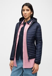 Γυναίκα που φοράει ένα καπιτονέ ναυτικό μπουφάν πάνω από μια ναυτική μπλούζα και ένα ροζ πουκάμισο από κορδόνι με κουμπιά, συνδυασμένα με ανοιχτόχρωμα μπλε τζιν, στέκεται μπροστά σε μια απλή επιφάνεια.