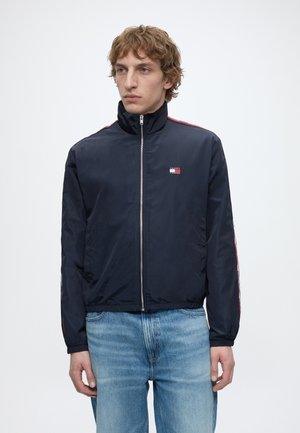 Ung mand med krøllet blondt hår iført en marineblå jakke med lynlås med røde striber og lyseblå jeans, stående mod en ensfarvet hvid baggrund.