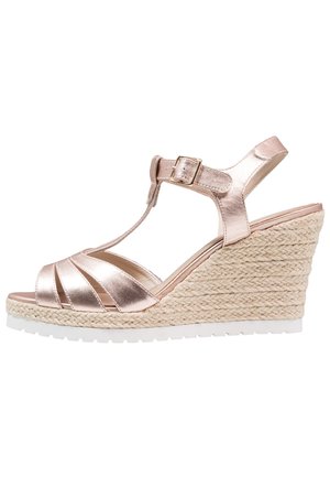 Metaliczne sandały na klinie w kolorze różowego złota z paskiem w kształcie litery T, obcas owinięty jutą i białą gumową podeszwą. Paski skrzyżowane dla lepszego wsparcia.