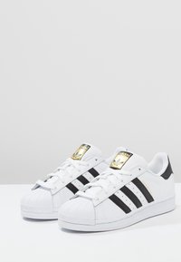 Vita Adidas Superstar-sneakers med svarta ränder och guldiga logotypdetaljer, visade på en vit yta mot en ljusgrå bakgrund.