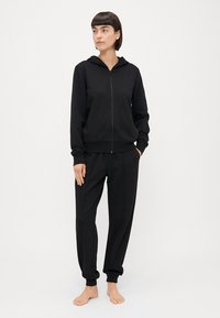 Crna zip-up kapuljača s suptilnim logotipom, uparena s odgovarajućim crnim trenirkama; obje od mekog materijala s rebrastim manžetama.