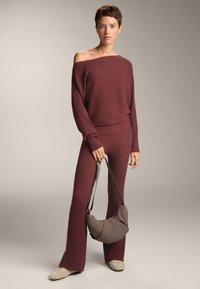 Rebrast pulover v maroon barvi z obliko brez ramenskega dela, v kombinaciji s usklajenimi širokimi hlačami. Drži rjavo torbico s podlogo; opazna teksturirana površina.