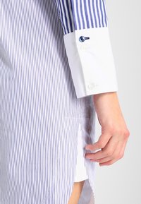Pánská košile s úzkými modro-bílými pruhy, bílý manžetový otvor s knoflíky a boční rozparek pro snadný pohyb. Hladká bavlněná textilie.