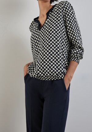 Woman wearing a black and white geometric patterned blouse with elastic hem and navy blue pants, hands in pockets against a plain background.