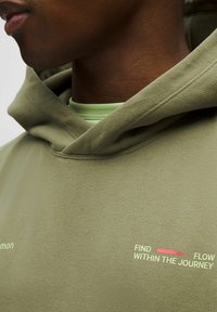 Hoodie vert olive en tissu doux, avec une capuche et des détails textuels à l'avant dans des couleurs contrastées. Superposé avec une chemise vert clair en dessous.