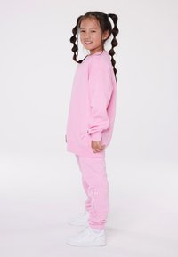 Rozā sporta krekls un atbilstoši bikses, kas izgatavotas no mīksta materiāla. Iezīmēta ar ribotām piedurknēm un potīšu maliņām, ar izšuvumu uz biksēm. Baltas sporta kurpes.