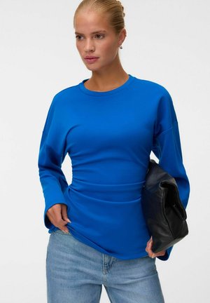 Femme aux cheveux blonds portant un haut à manches longues bleu vif et un jean bleu clair, tenant un sac en cuir noir plié sous le bras.