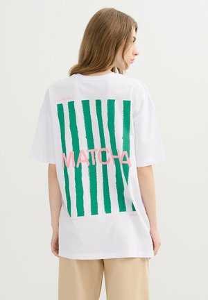Joven mujer con cabello largo castaño claro que lleva una camiseta blanca extragrande con rayas verticales verdes y la palabra "MATCHA" en rosa en la parte de atrás.