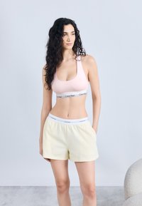 Γυναίκα με μακριά σγουρά μαλλιά, που φοράει ανοιχτό ροζ αθλητικό σουτιέν και ανοιχτό κίτρινο σορτς με ζώνες στη μέση με την επωνυμία Calvin Klein, στέκεται με το ένα χέρι στην τσέπη.