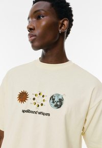 Helle gelbe Baumwoll-T-Shirt mit einem Grafikdesign von einer Sonne, einem Mond und Planeten, begleitet von dem Text "zauberhafte Flüstern."