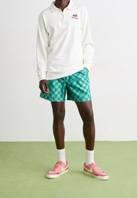 Polo blanca de manga larga con cremallera y logo de Nike, combinada con shorts de cuadros verdes. Zapatillas rosas con detalles blancos y calcetines.