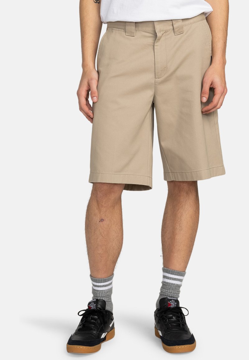 Pantaloni corti khaki con taglio dritto, passanti per cintura e design frontale piatto. Abbinati a sneakers nere e calze grigie a righe.