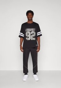 Άντρας που στέκεται μπροστά σε απλή τοιχογραφία, φορώντας μαύρη μπλούζα "Champion 92", μαύρο παντελόνι με λευκές ρίγες και λευκά αθλητικά παπούτσια.